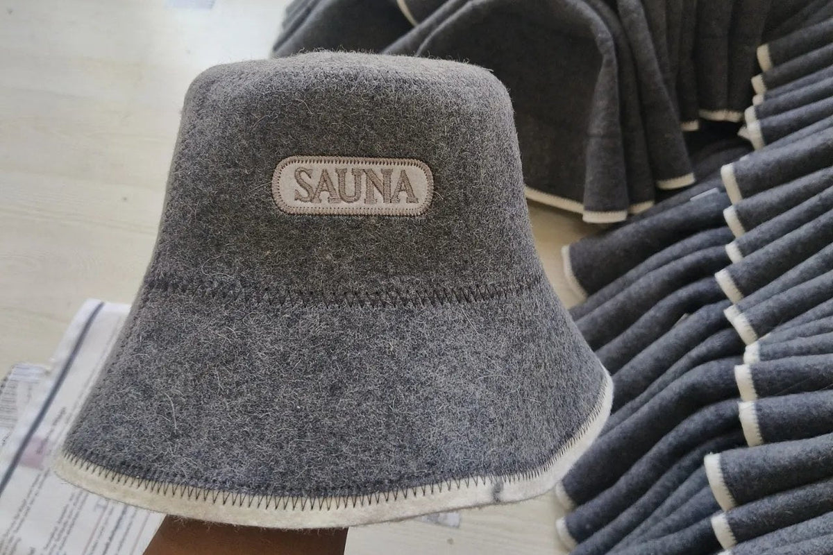 Grauer Saunahut aus Filz mit der Aufschrift „Sauna“ und hellem Rand, in der Hand gehalten, dahinter weitere Saunahüte